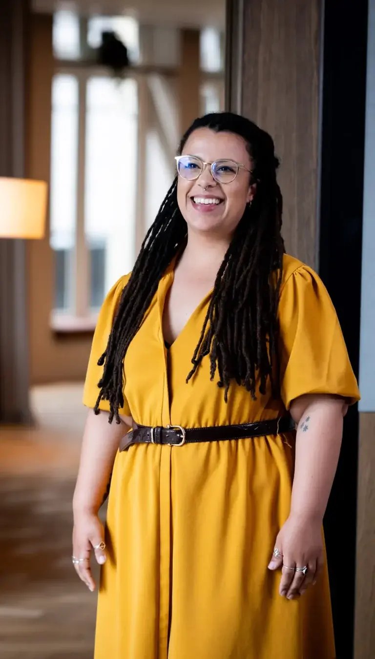 Smiling woman in a mustard yellow dress standing in a modern office environment, representing the human side of Nétive VMS.