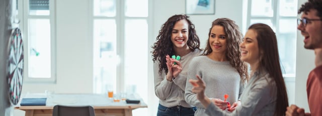 Vier collega's lachen en spelen darts samen in een informele kantoorruimte.