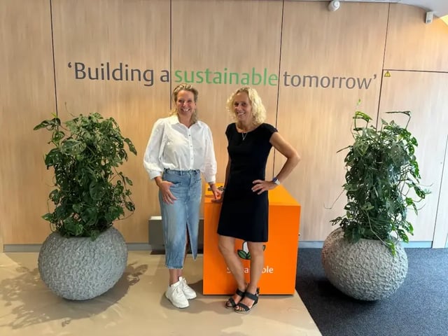 Twee vrouwen, vermoedelijk Jolanda en Meke, staan lachend naast elkaar in een kantoorruimte met de tekst "Building a sustainable tomorrow" op de muur.