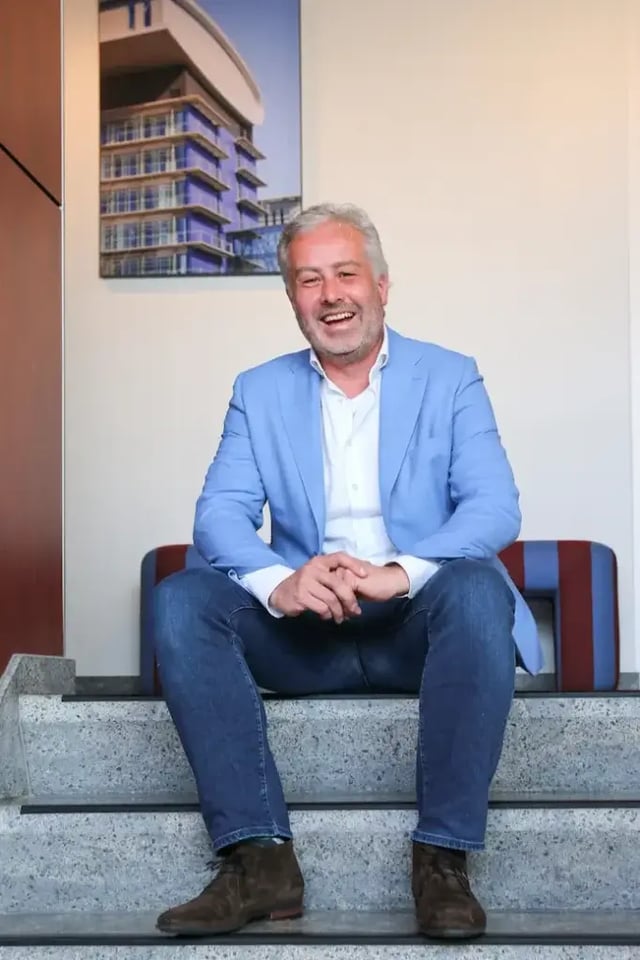 Man in lichtblauw colbert zit lachend op een trap met een modern gebouw op de achtergrond aan de muur.