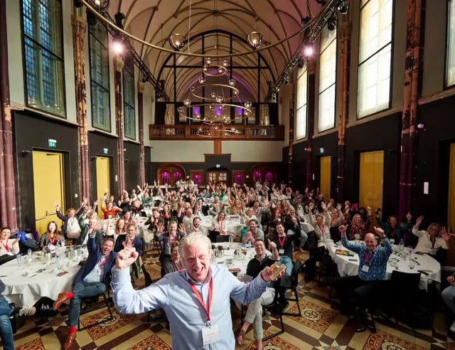 Man maakt een selfie met een enthousiast publiek in een grote, sfeervolle zaal met hoge ramen en sierlijke verlichting.