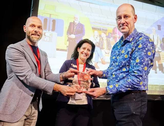 Drie mensen ontvangen samen de TTM Award 2025 voor NS op een podium met een foto van een trein op de achtergrond.
