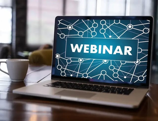 Laptop op een tafel met het woord "WEBINAR" op het scherm en een koffiekopje ernaast.