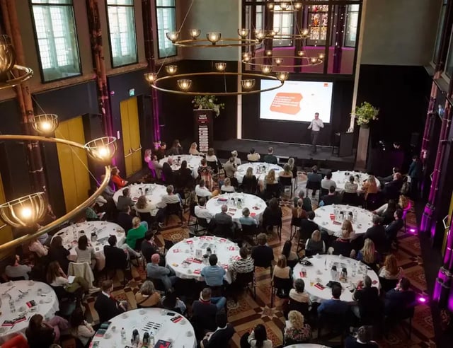 Overzicht van een conferentiezaal van bovenaf met ronde tafels en publiek gericht op een spreker op het podium.