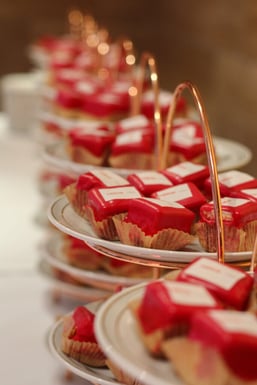 Etagères met roodglazuur petit fours in papieren vormpjes, netjes uitgestald op een witte tafel.